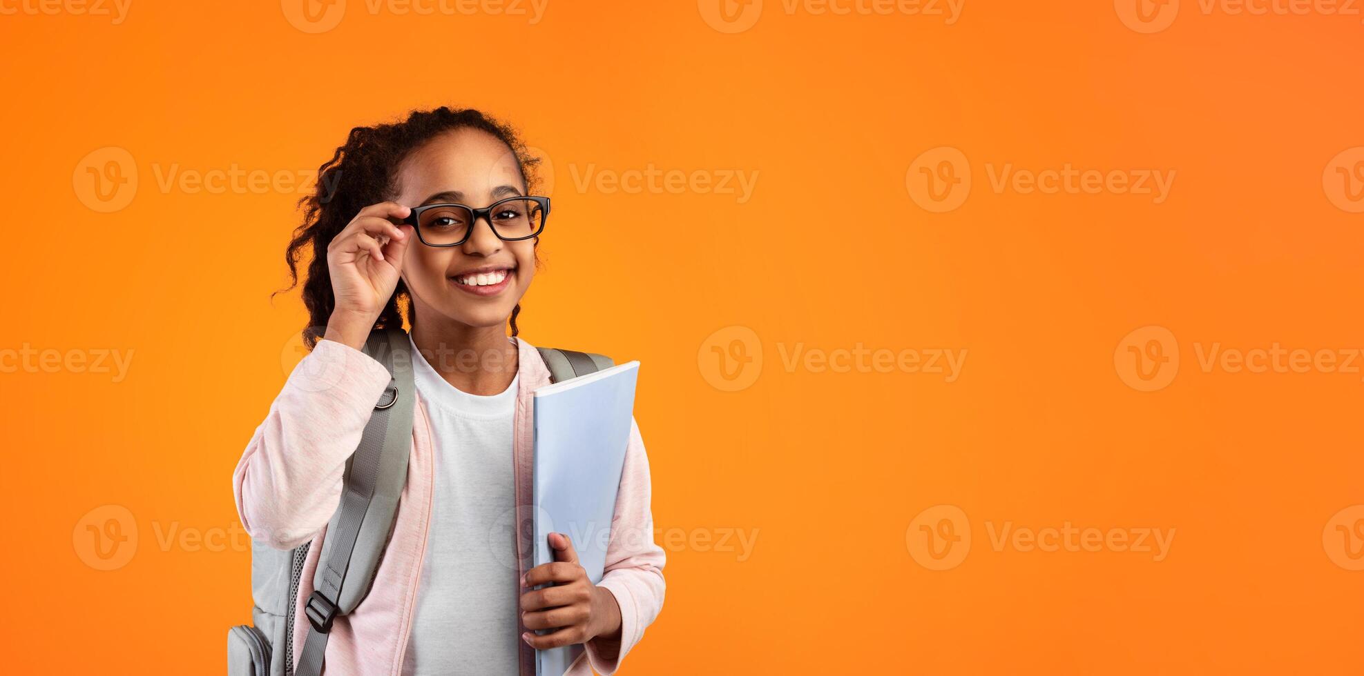 Education Concept. Portrait of positive smiling African American girl touching glasses holding textbooks, wearing backpack and looking at camera isolated on yellow studio background photo