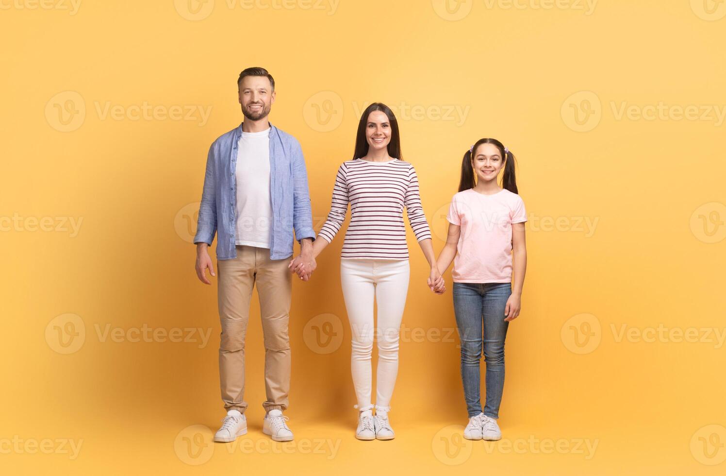 A loving family of three, consisting of a father, mother, and their teenage daughter, holds hands while standing together against a bright yellow backdrop, showcasing their bond and affection. photo