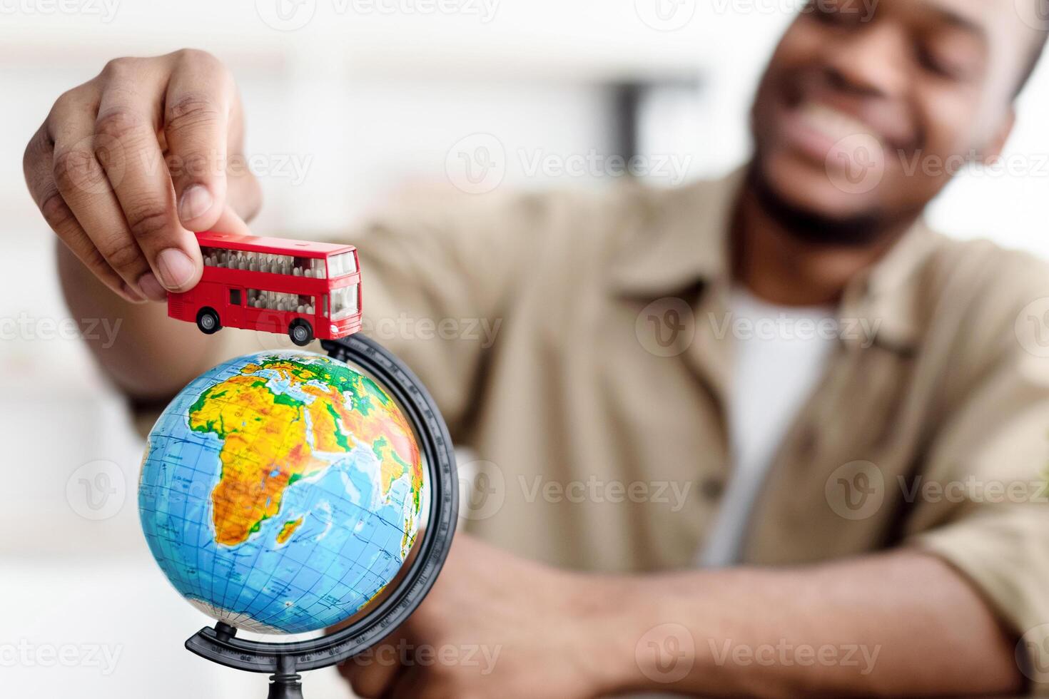 African American Male Tourist Planning Vacation Playing With Red Bus Model And World Globe Sitting At Desk Indoor. Man Dreaming About Trip To Great Britain. Selective Focus On Toy photo
