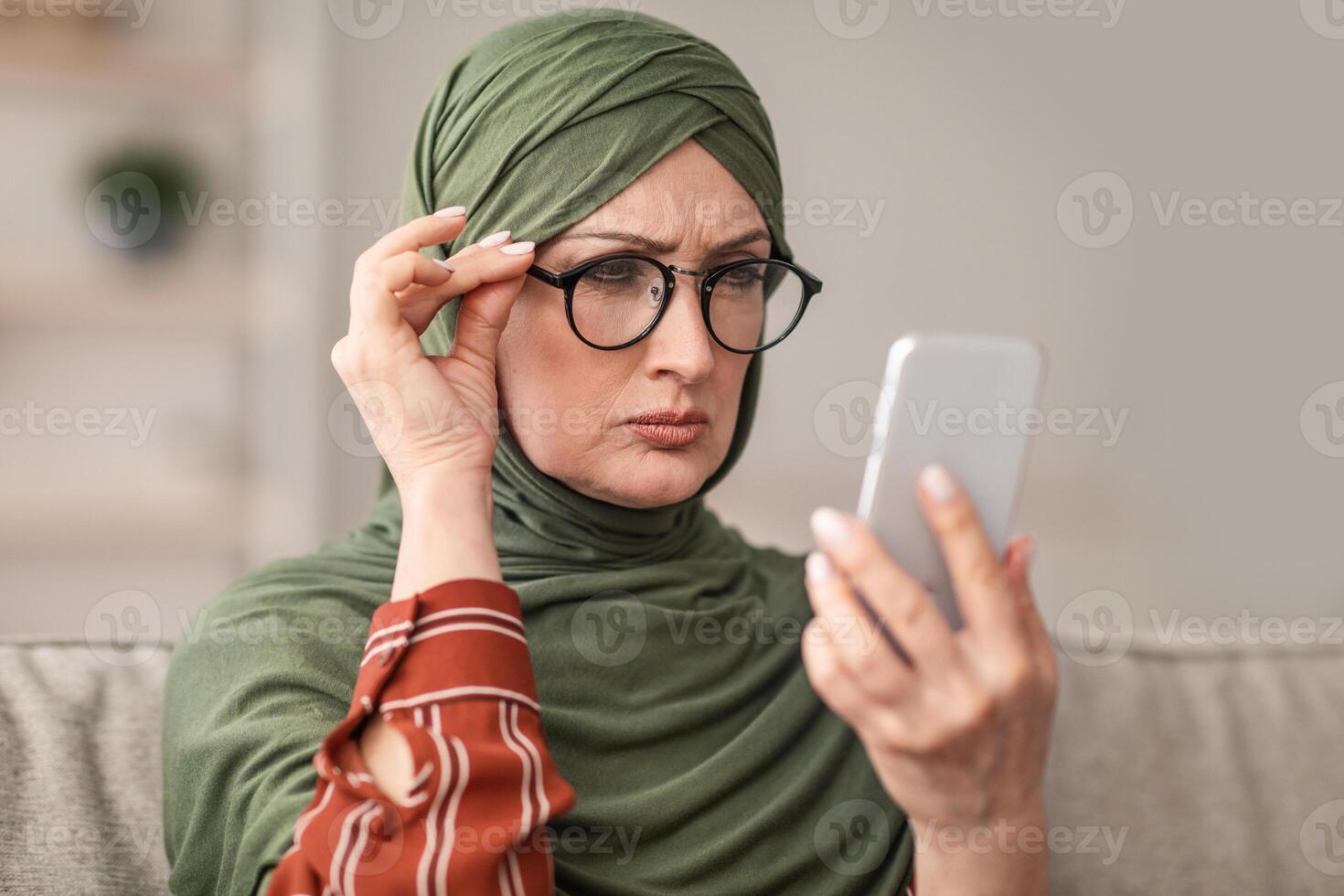 Poor Eyesight. Senior Middle-Eastern Woman In Hijab Squinting Eyes Reading Message On Phone, Wearing Eyeglasses Sitting At Home. Mature Muslim Female Having Bad Vision Health Problems photo