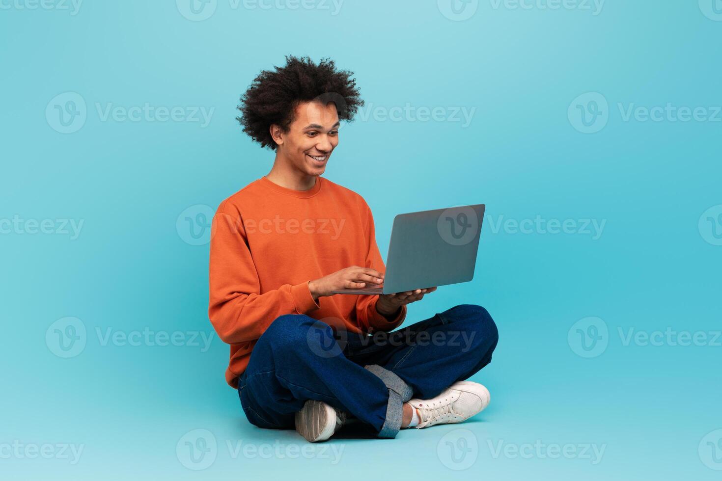 A joyful African American man with curly hair sits cross-legged, engaging with his laptop. He wears an orange sweater and looks delighted in a bright, minimalist environment. photo