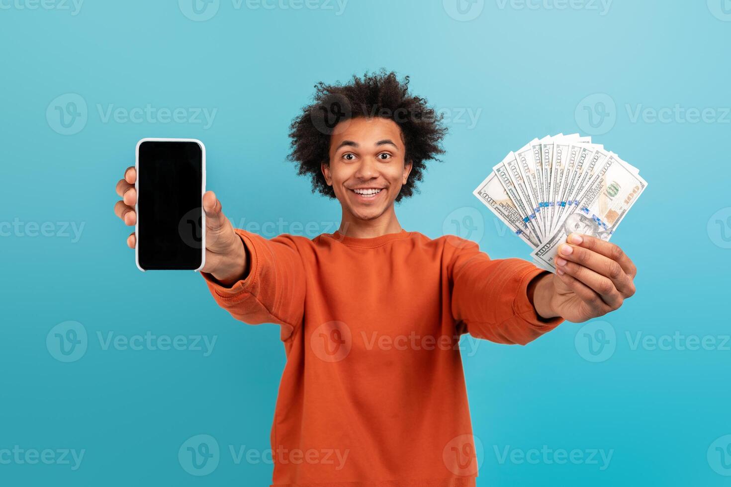 A young African American man with curly hair smiles widely while holding a smartphone in one hand and a stack of cash in the other, embodying a cheerful lifestyle and achievement. photo