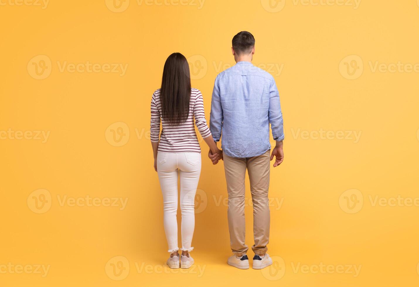 A Caucasian couple stands together, holding hands and facing away. They express love and affection while dressed casually, isolated against a vibrant yellow background. photo