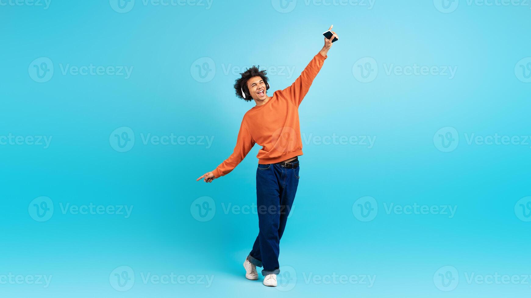 A young black man in a vibrant orange sweater playfully poses against a blue background. He holds a smartphone while expressing happiness and freedom, embodying a lively lifestyle. photo