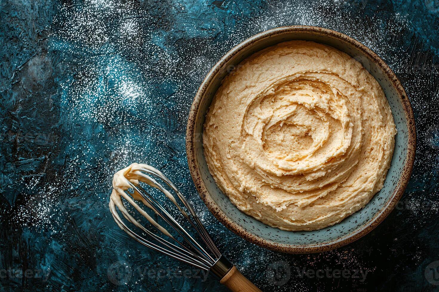 Smooth and creamy buttercream frosting is blended in a bowl, with a whisk resting beside it. The dark blue surface adds contrast, making the frosting the focal point of the setup. photo