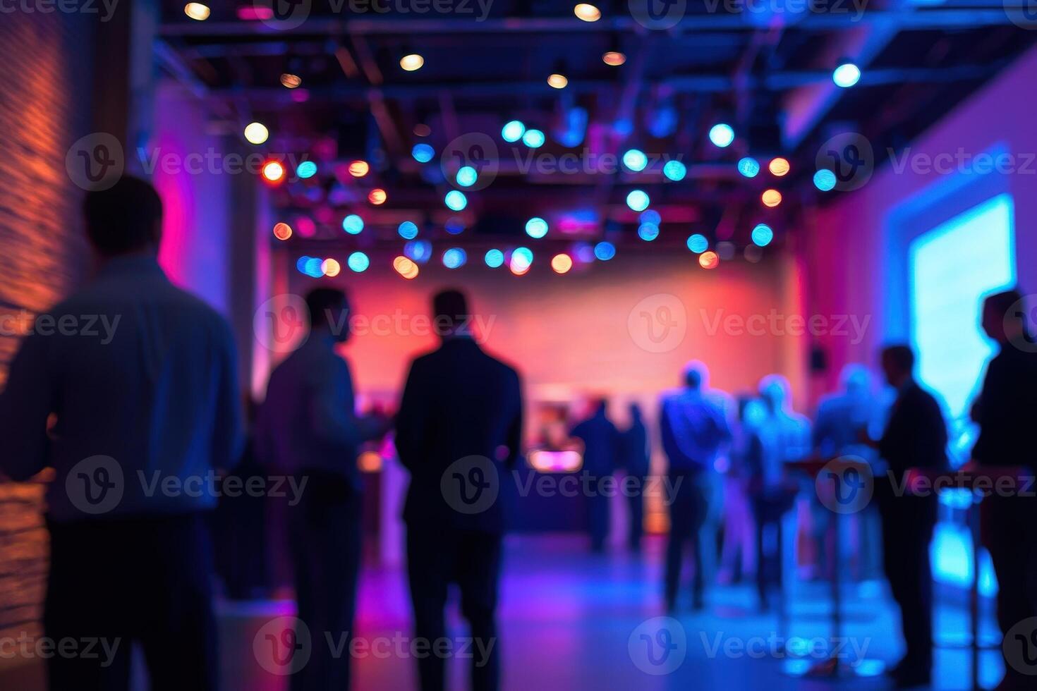 Attendees gather at a contemporary venue featuring vibrant lighting for a networking event. Individuals converse and connect, creating a lively atmosphere in the dimly lit space. photo