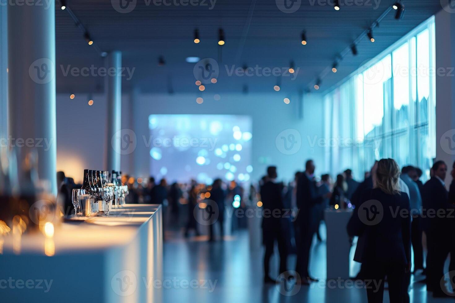 A sophisticated gathering takes place in a contemporary venue. Guests mingle and enjoy drinks while a large projection adds to the ambiance, creating a lively atmosphere for conversation. photo