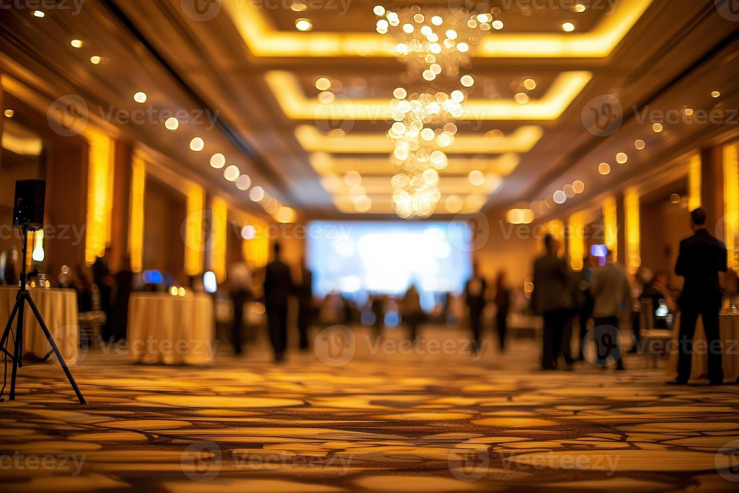 A spacious banquet hall is softly lit with golden tones, featuring elegantly dressed guests mingling. The atmosphere is festive as a large screen displays visuals, creating an inviting ambiance. photo