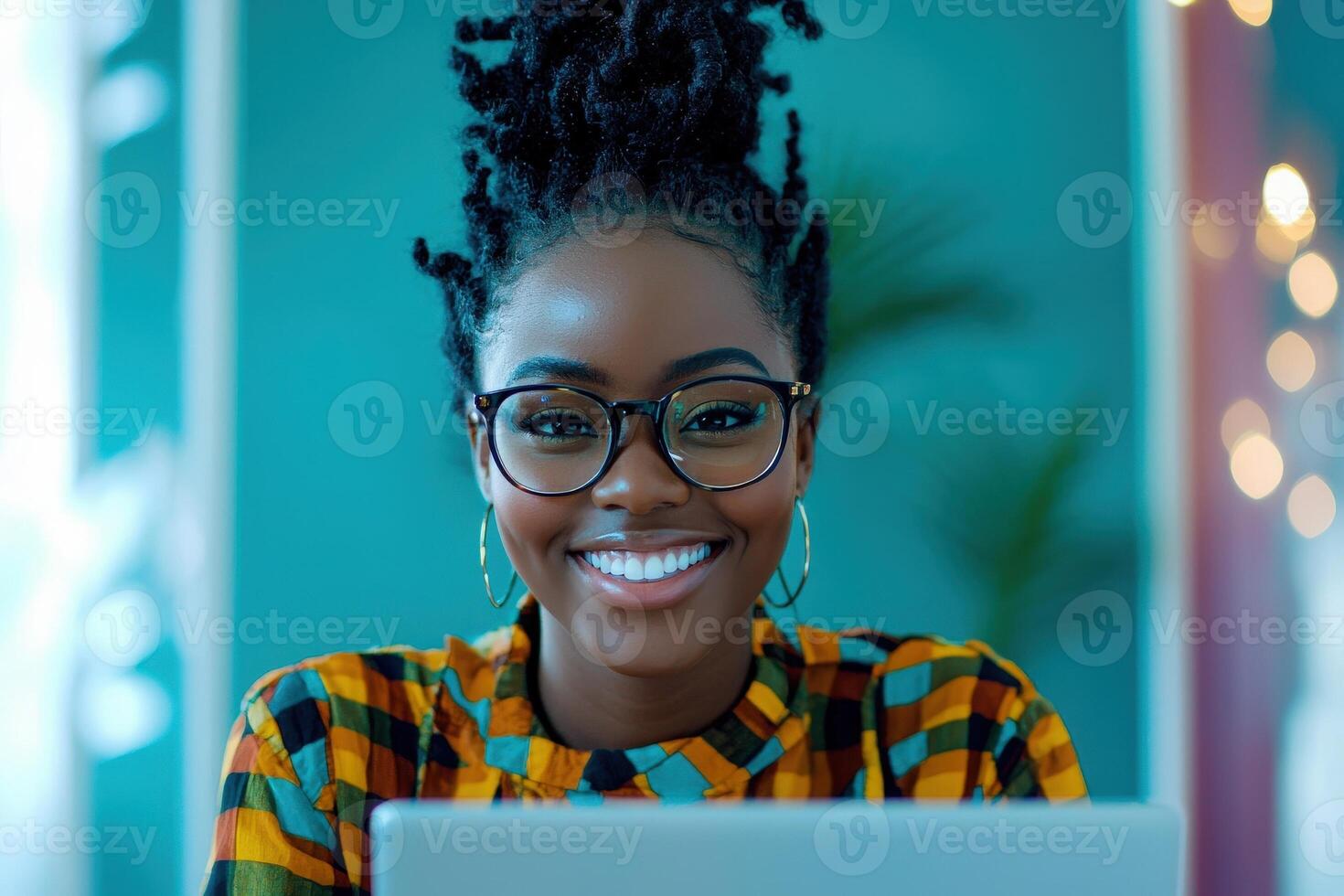 A young woman with natural hair smiles brightly while sitting at a desk in a colorful room. She is wearing glasses and a patterned shirt, focused on her laptop against a lively background. photo