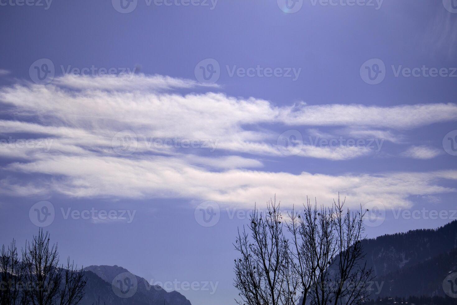 Cirrus Clouds Streaking Across a Blue Sky Above Mountain Peaks Bare Tree in the Foreground photo