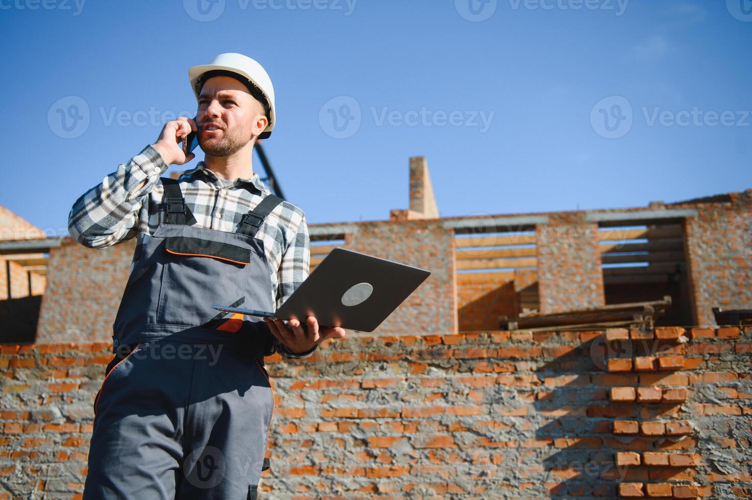 Engineer, bearded man wears safety helmet inspecting housing development project at construction site using laptop computer. Contractor manager examining residential building estate infrastructure photo