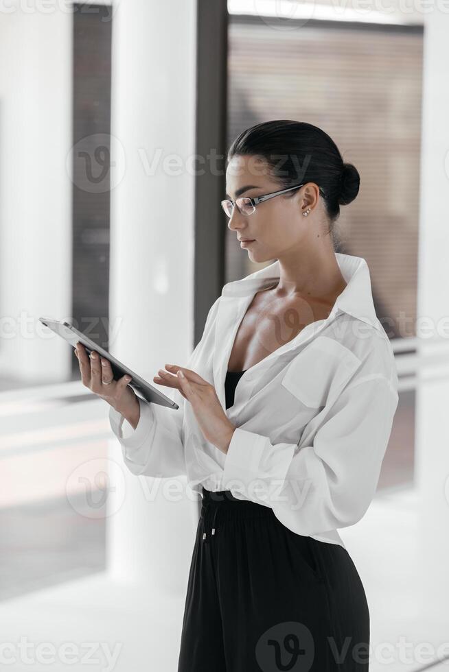 Young focused intern, student holding electronic gadget in office, studying educational app on pad, reading guidance. Manager sharing content on digital device. Young multi-ethnic female in formalwear photo