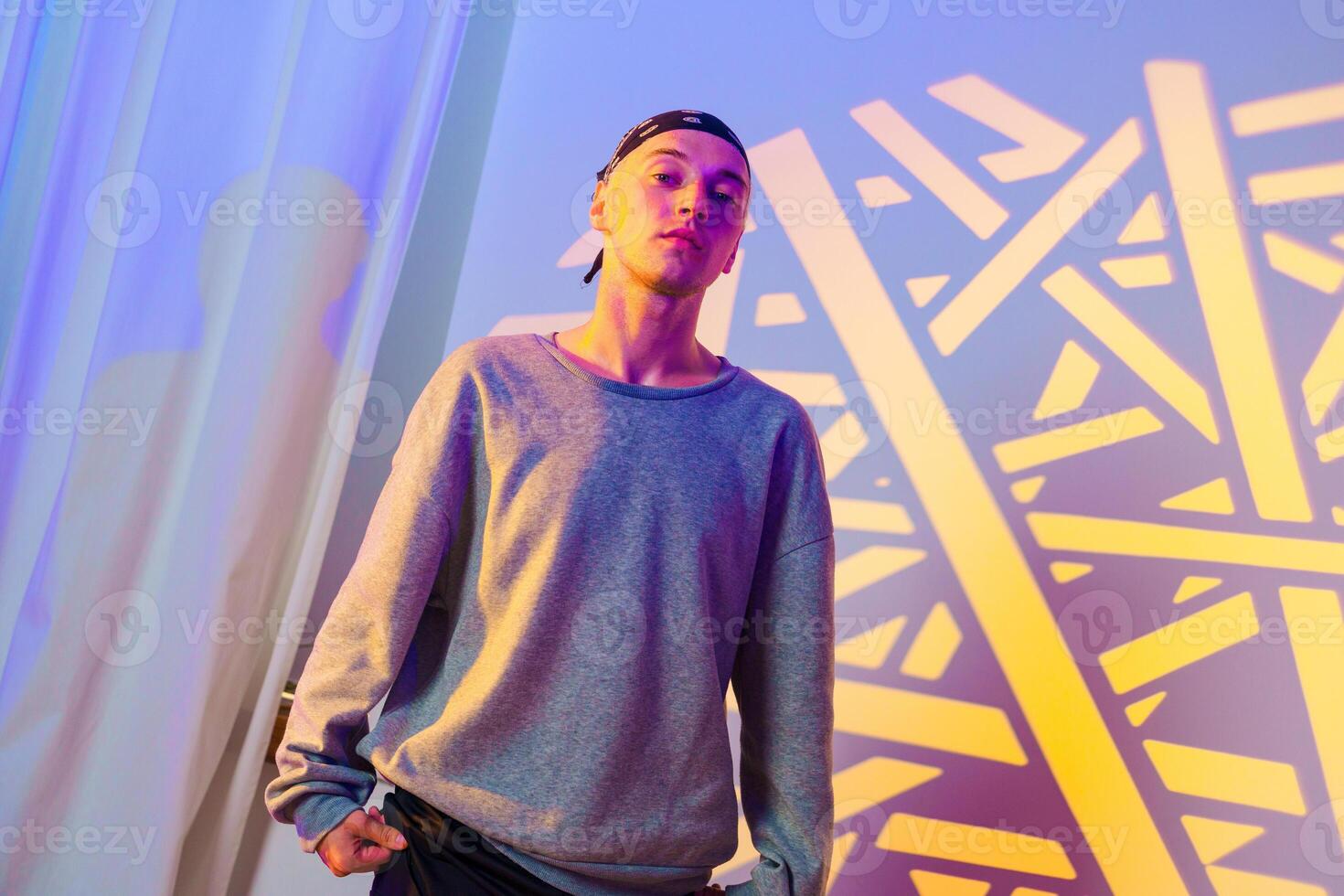 A young man in a gray sweater stands in front of a colorful background, looking directly at the camera photo