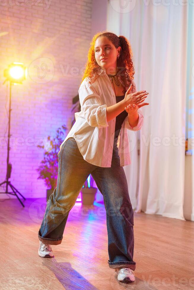 A young woman with curly red hair dances in a studio, wearing a white shirt and blue jeans photo