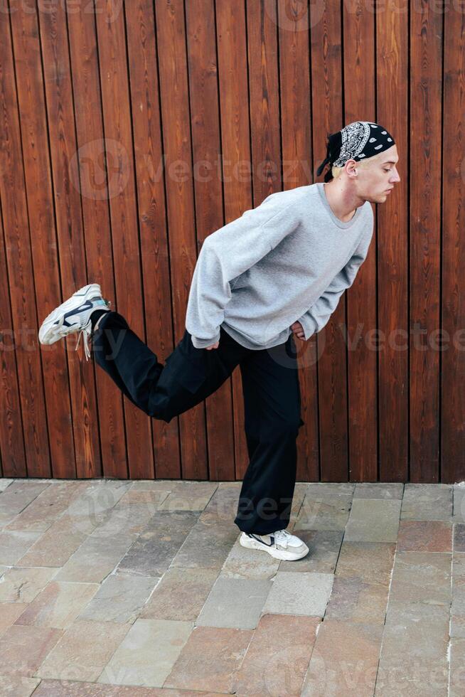 A man in a grey sweater and black pants is doing a dance move in front of a wooden wall photo