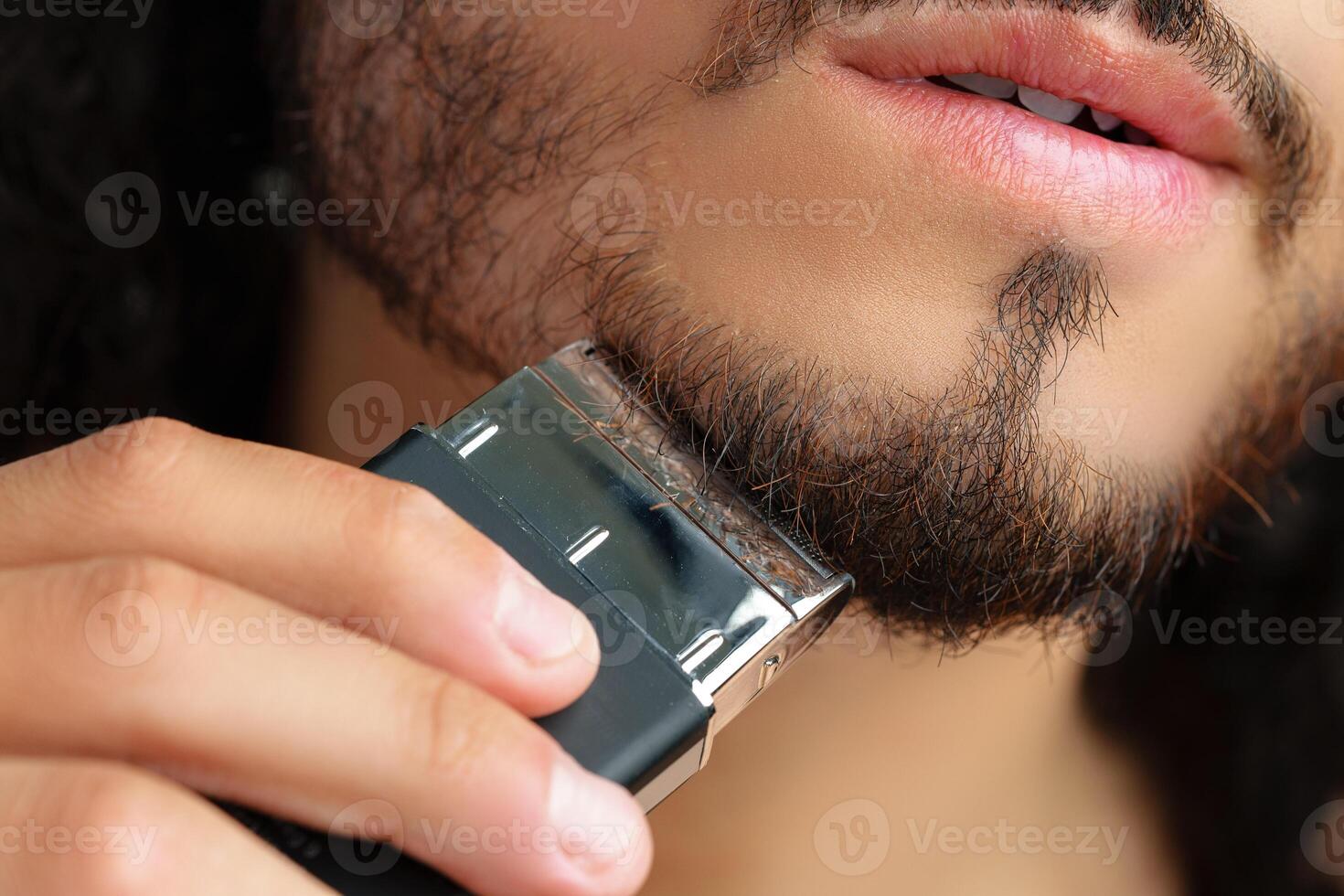 Man trimming his beard indoors using an electric trimmer while showcasing his personal grooming routine photo