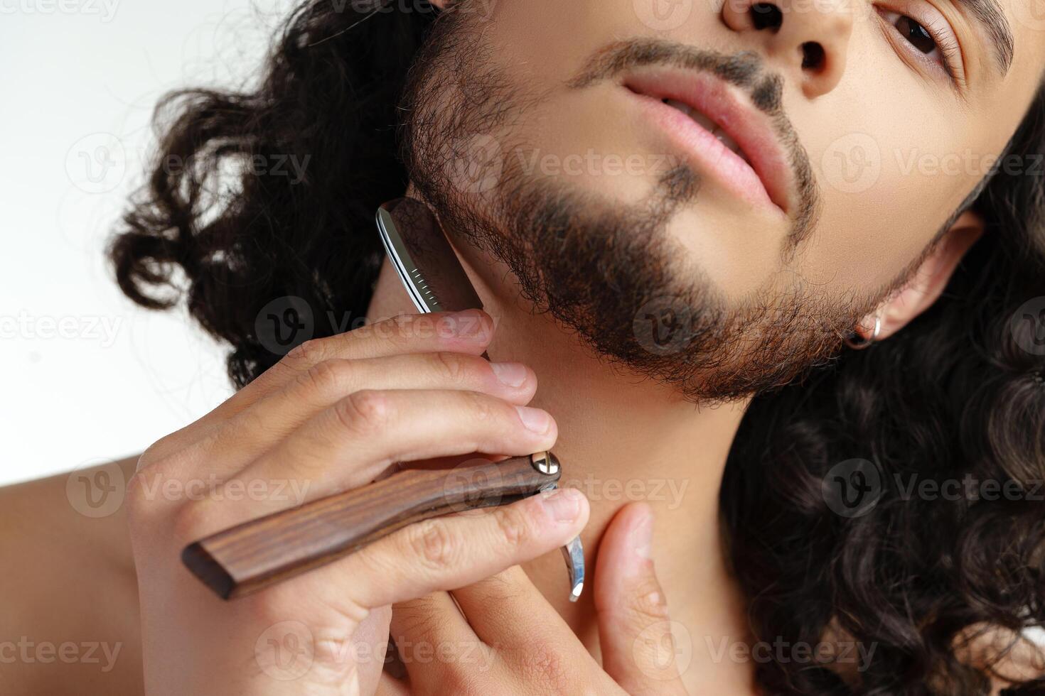 Man with curly hair grooming his beard carefully using a straight razor against a plain white background photo