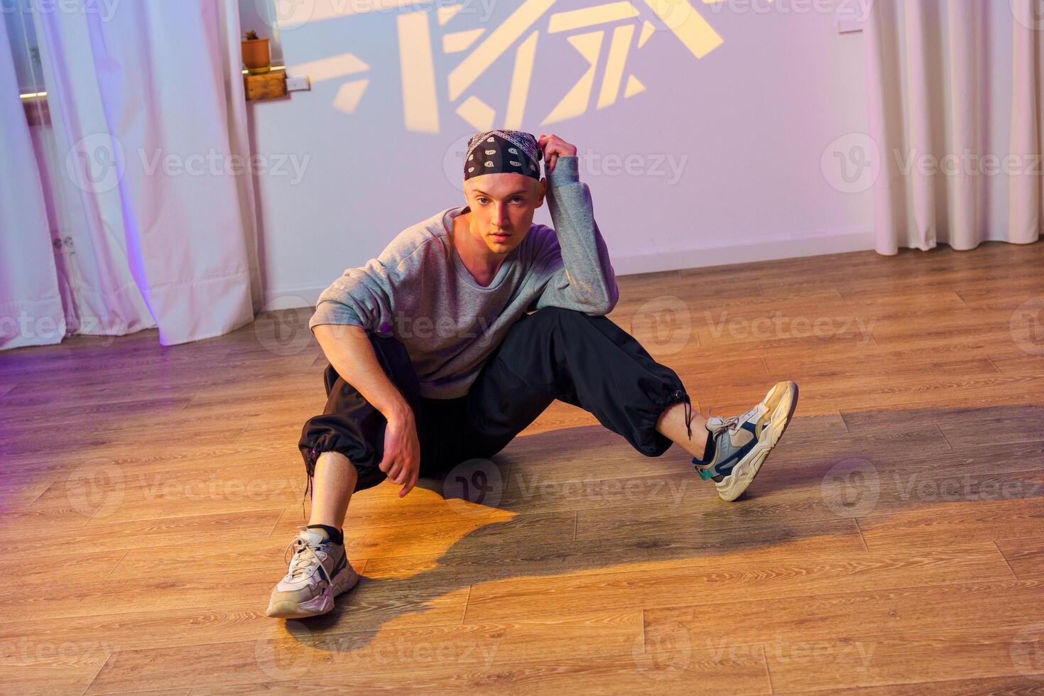 A young man sits on a wood floor in a dance studio, wearing a bandana and sneakers photo