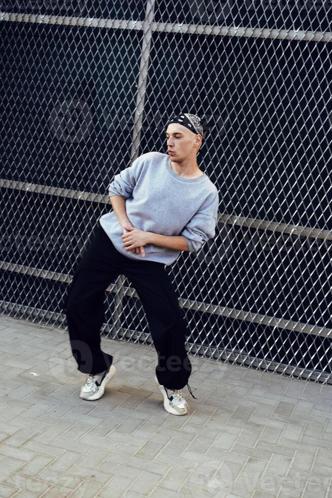 A man dances in front of a chain link fence, wearing a grey sweatshirt, black pants, and white sneakers photo