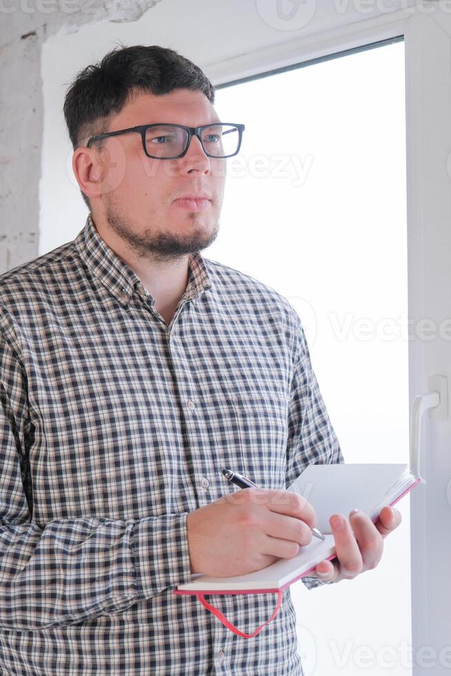 man in plaid shirt sitting at windowsill , thinking and writing in a notebook photo