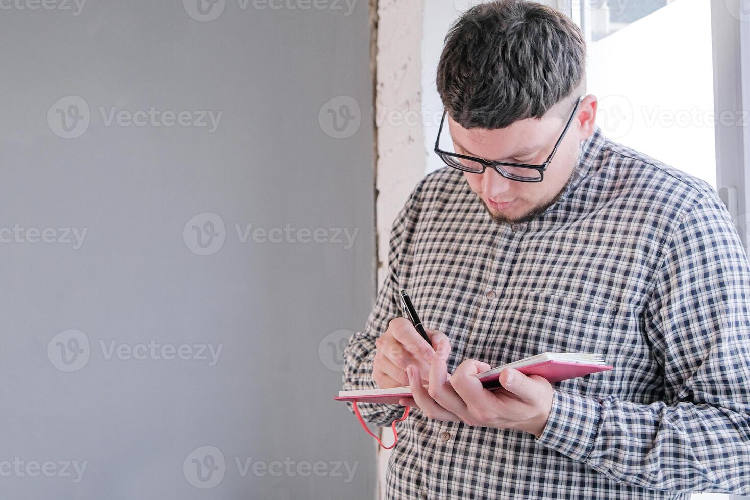 man in plaid shirt sitting at windowsill , thinking and writing in a notebook photo