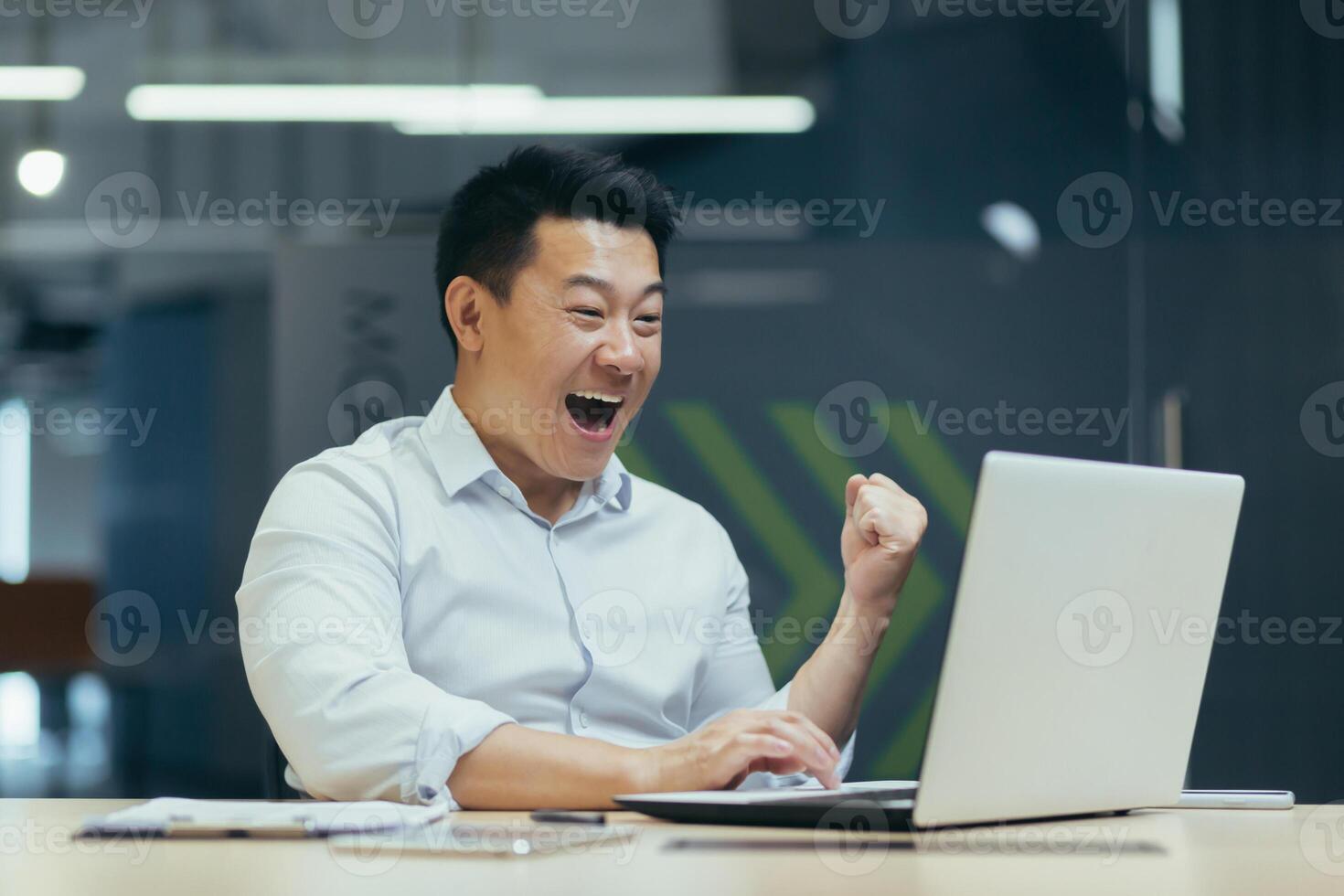 Young man businessman freelancer Asian looks at the laptop monitor in the office, received important good news, rejoices, points with his hand yes, shouts with happiness. photo