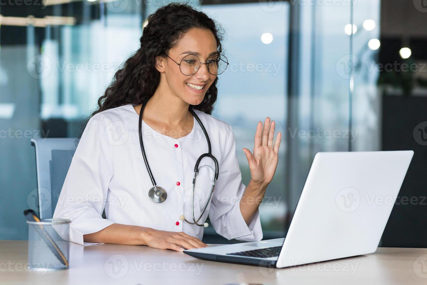 The doctor is online. Young Latin American female doctor consults online patients via connection from a laptop. She is sitting in the office in a medical gown. He waves at the camera, smiles. photo