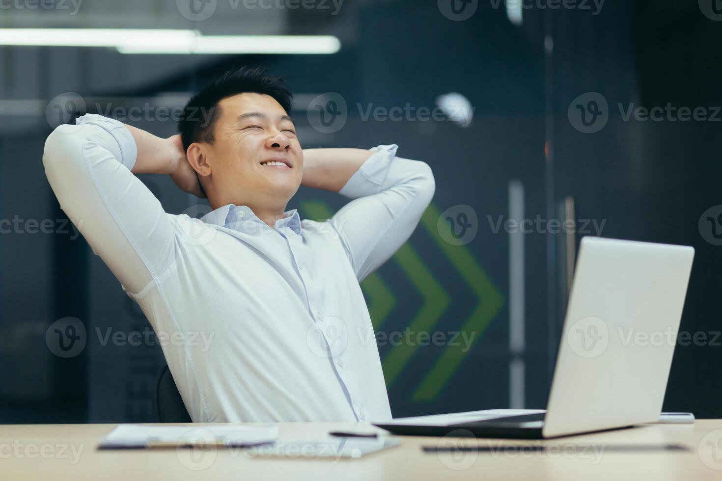 A break at work. A young Asian man is resting at the workplace at the desk in the office. He sits with his hands behind his head, closes his eyes, rests. photo