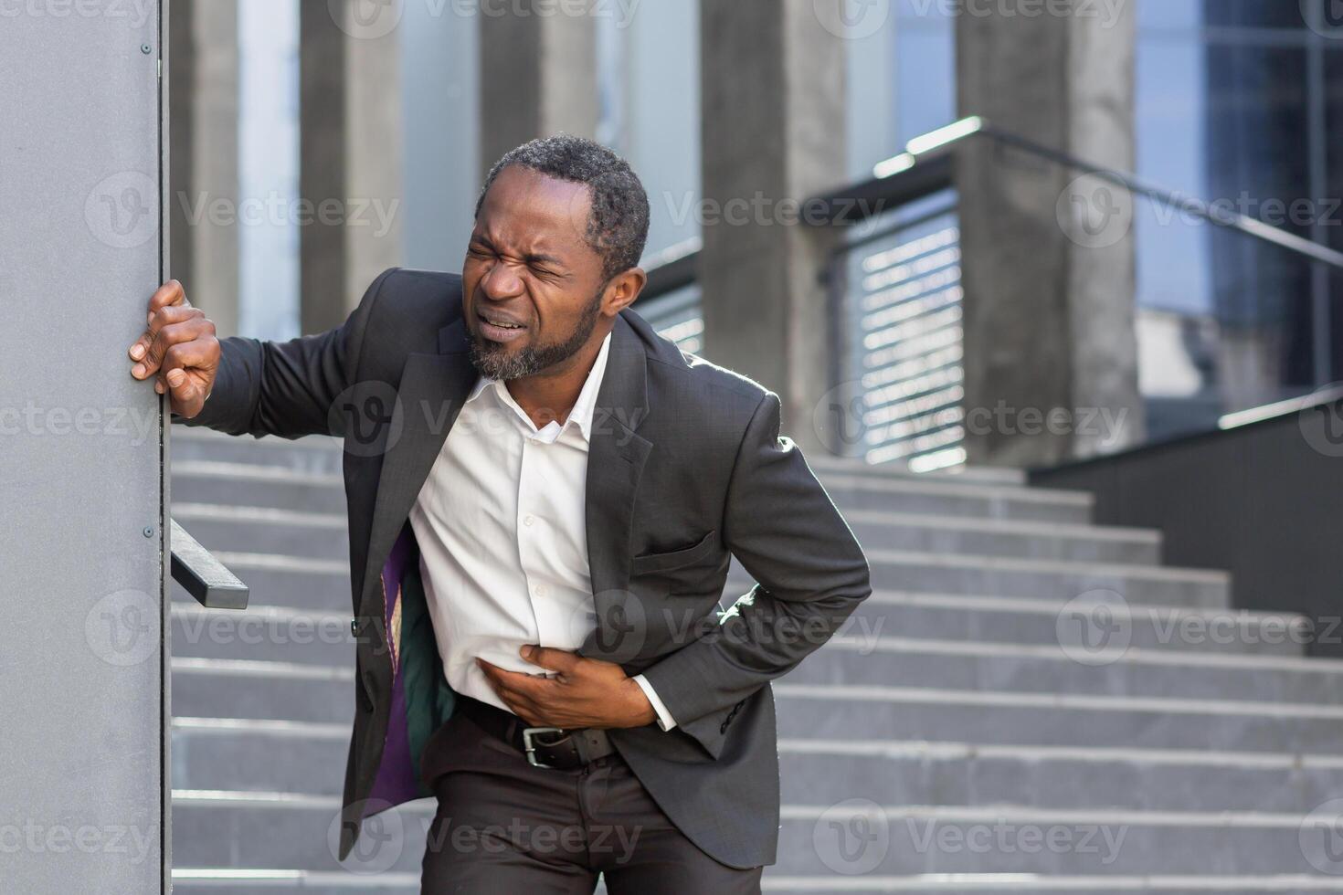 An African American man in a suit has a stomach ache outside an office. He holds his stomach, feels severe pain, upset stomach, poisoning, diarrhea, hunger. Contorted in pain, holding on to the wall. photo