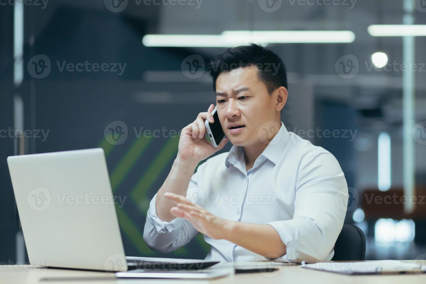 Worried and nervous young Asian man talking on the phone in the office, looking at the laptop, solving problems, questions. photo