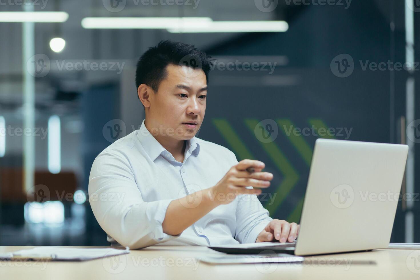 Business online meeting. A young asian serious businessman holds a meeting with partners, clients, a conference. Sitting at the desk in the office, looking at the laptop, explaining, smiling. photo