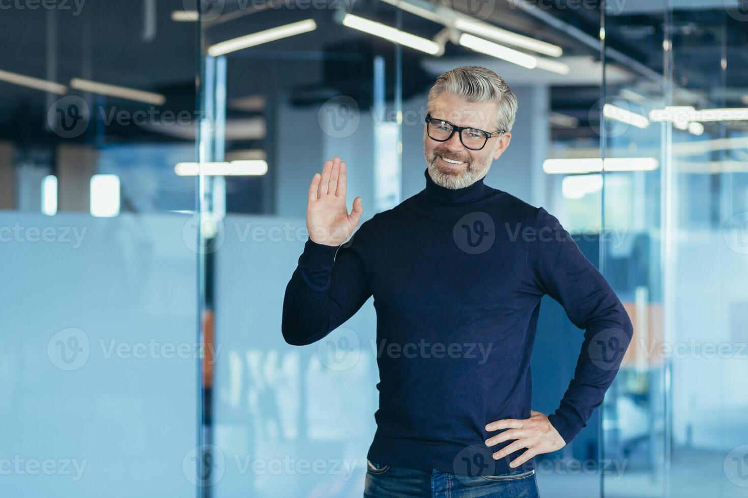 Portrait of successful financier in modern office, man looking at camera and smiling holding hand up greeting gesture, gray haired senior experienced businessman with beard and glasses, investor owner photo