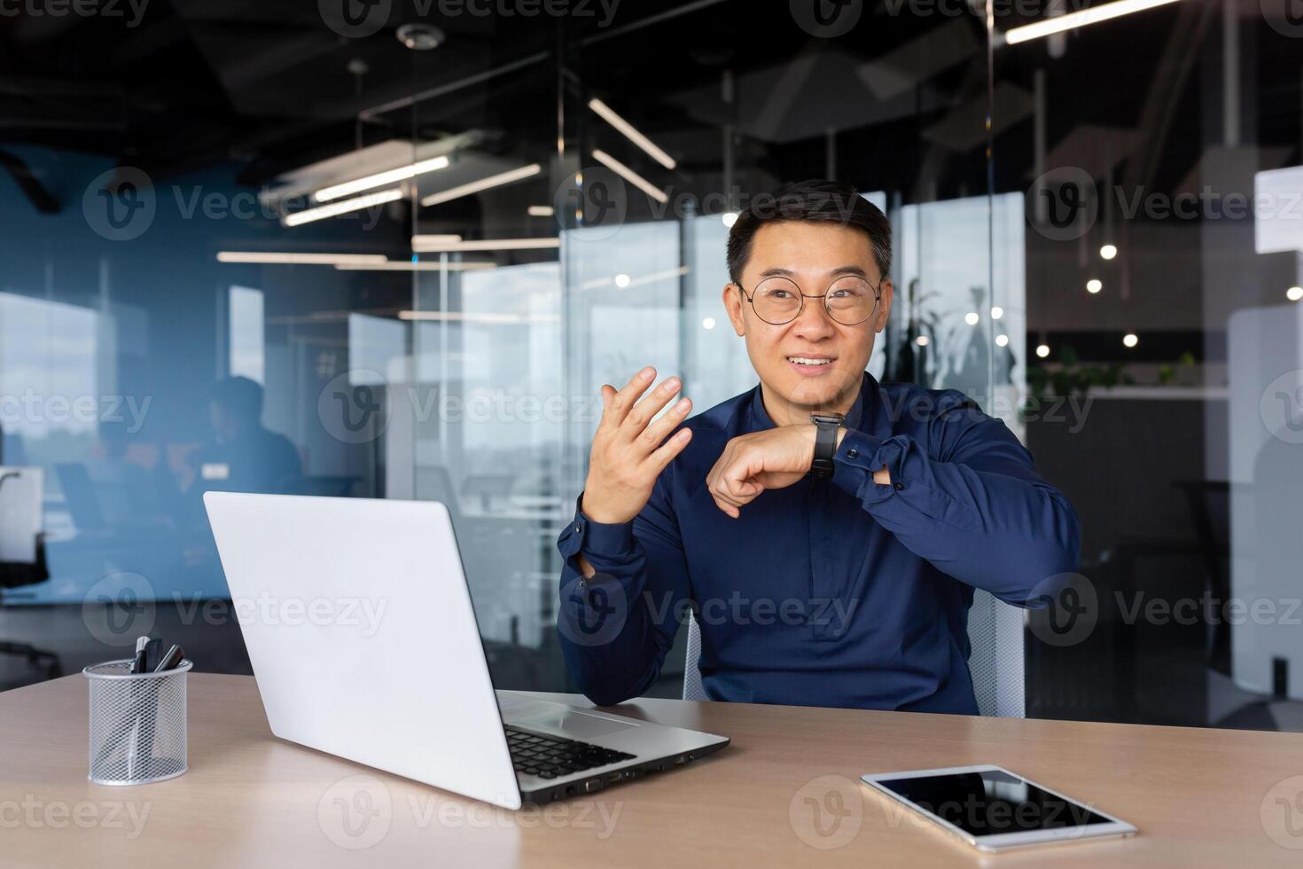 Asian businessman uses smartwatch to communicate, man gives commands to artificial intelligence using wrist watch, boss in shirt works inside office using laptop. photo