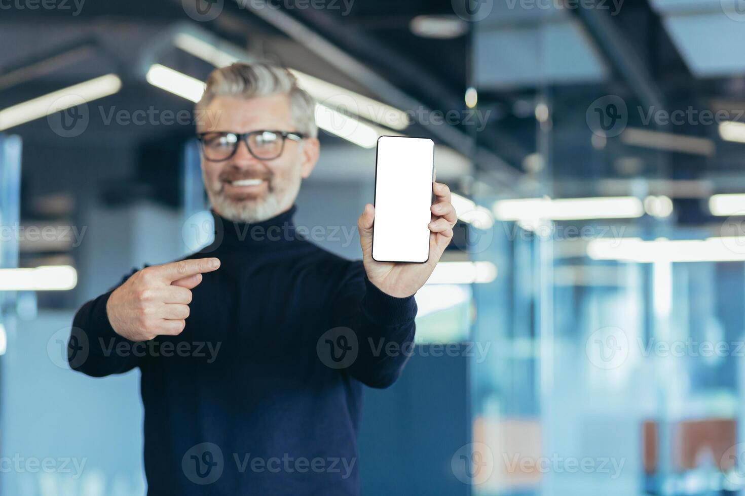 Senior successful gray-haired businessman in office, showing phone screen to camera with white screen background, man pointing with finger on copy space photo