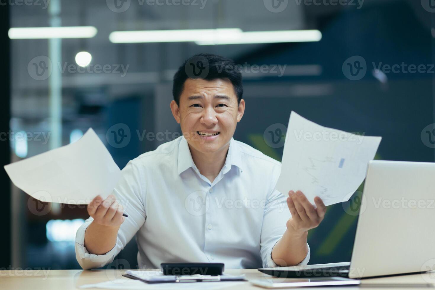 A worried young man, an Asian businessman, is sitting in the office at the table, holding documents in his hands. Got bad news, bills, financial problems, failed deal. photo
