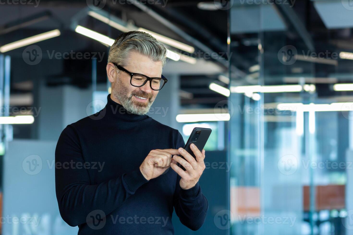 Mature adult man inside office standing near window, businessman typing message on phone and browsing internet pages, gray-haired boss smiling reading news online. photo