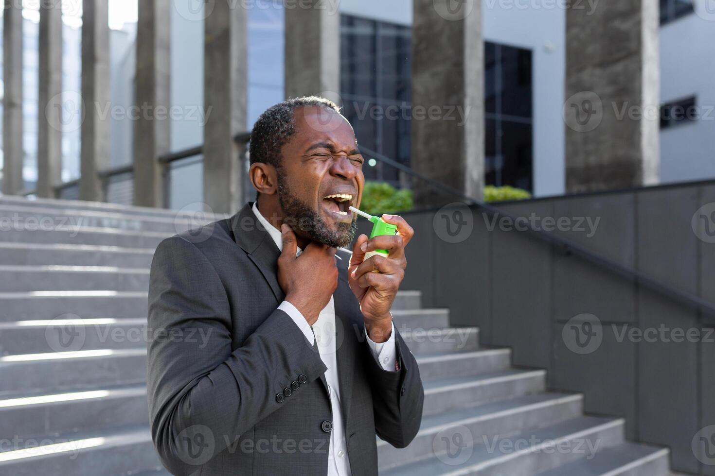 An African-American businessman is standing on the street near an office center and blowing an aerosol into his throat. He feels severe pain in the throat, cough, allergy, suffocation. photo
