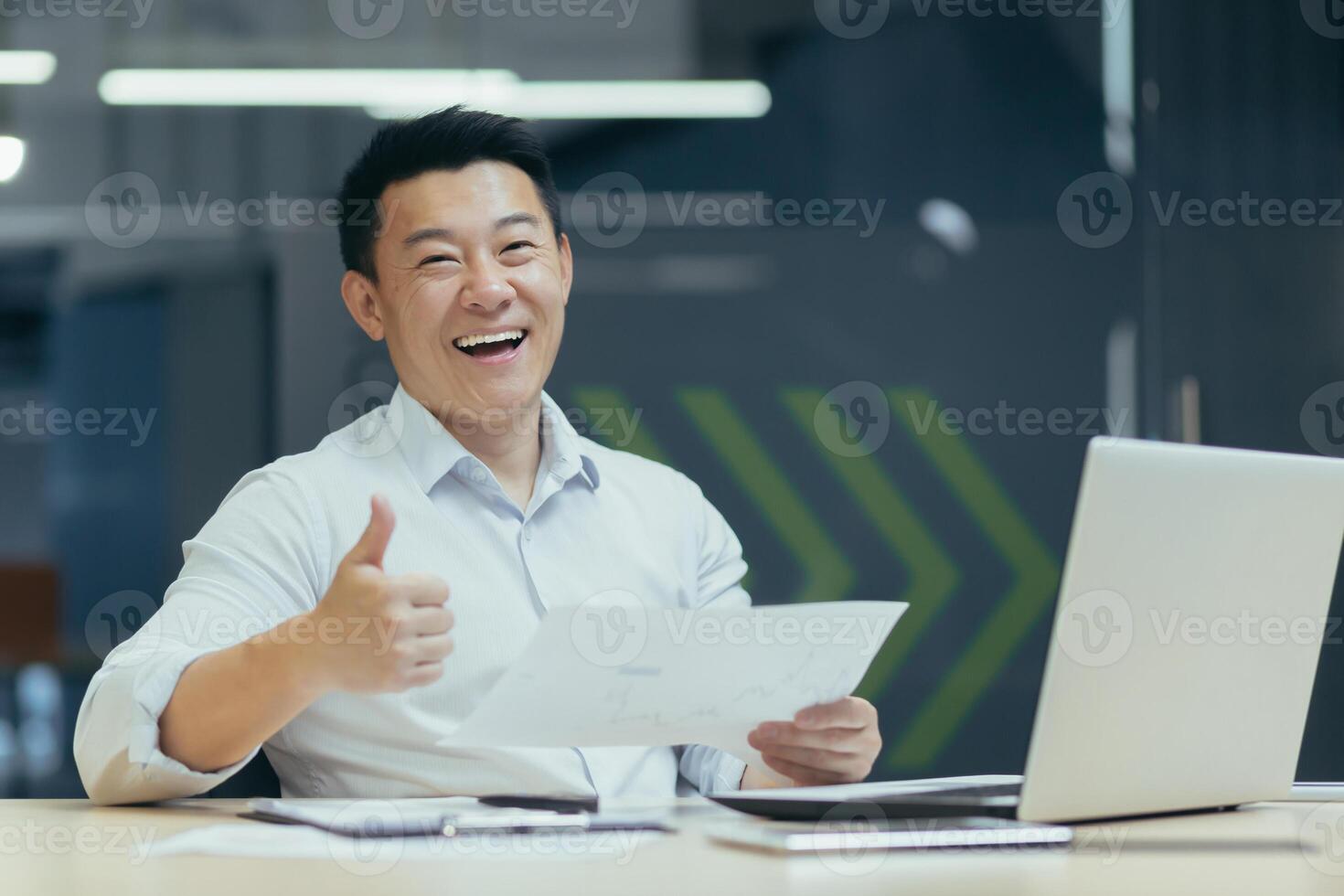 Portrait of a young Asian businessman man sitting at a desk in the office with a laptop and holding documents. rejoices in a successful deal and increased income. Pointing finger at camera super photo