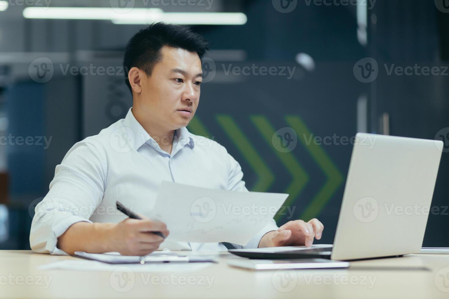 Portrait of young serious and focused Asian businessman working with documents on laptop. Sitting at a desk with a laptop in a modern office in a white shirt photo