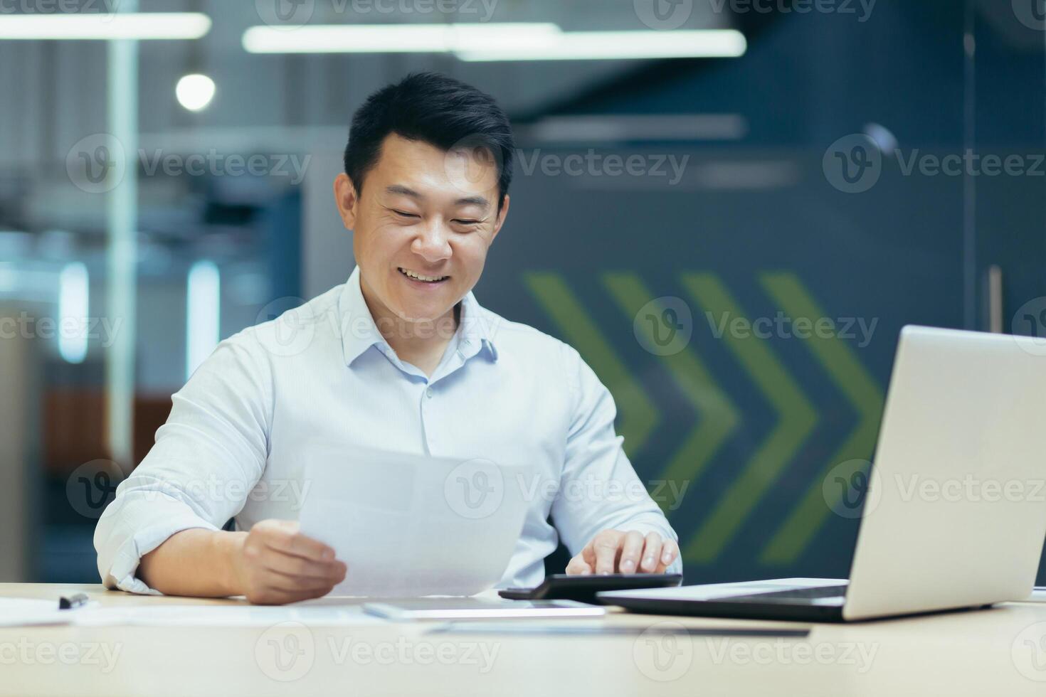 Portrait of a young Asian male accountant sitting at a desk in the office, holding documents in his hands, using a calculator. Works with accounts, budget, loans. photo