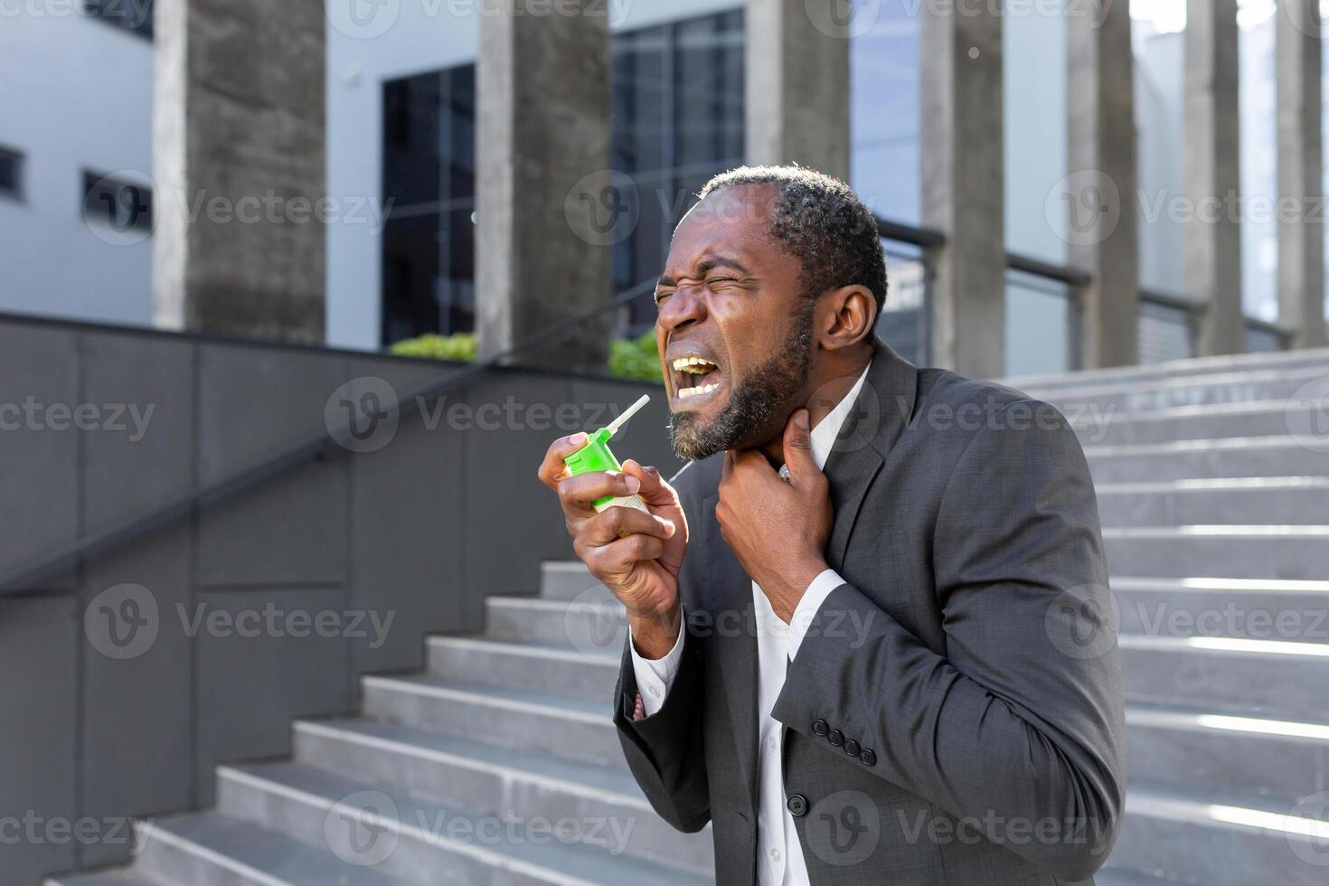 An older African American man in a suit clears his throat from pain and inflammation while standing outside an office center. photo