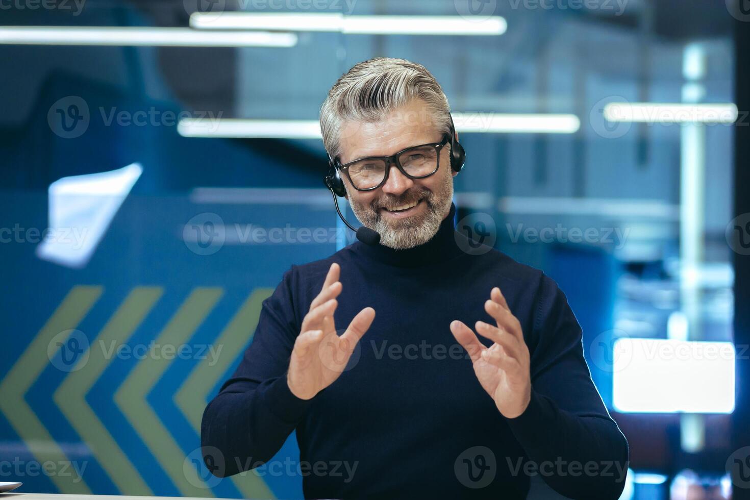 Senior gray haired businessman talking on call, using headset investor in glasses smiling and looking at camera, webcam view man working inside modern office building. photo