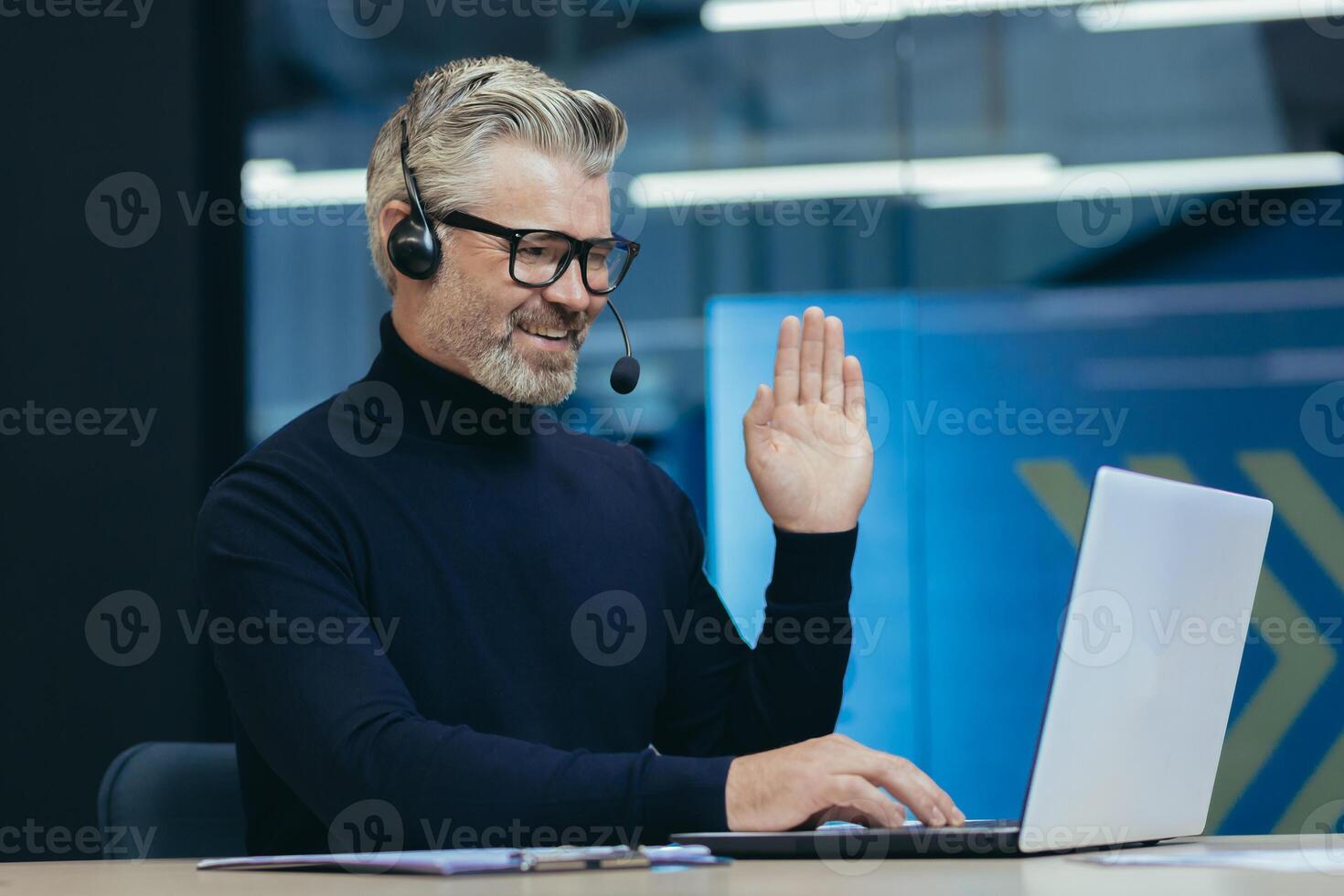 Online meeting with colleagues, senior gray-haired man working in modern office interior, executive with headset and glasses smiling and looking at laptop camera waving greeting gesture, call photo