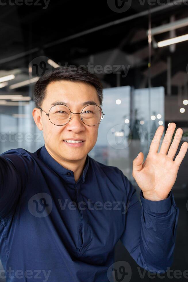 Vertical shot, Asian businessman talking on call, man looking at smartphone camera and waving cheerfully, greeting gesture, programmer working inside office in glasses and shirt. photo