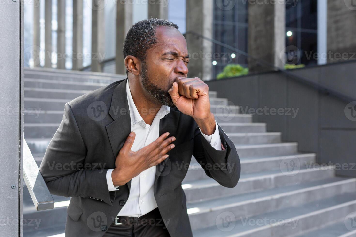 Cough attack on the street. Senior African American businessman standing and coughing suffocatingly, choking, covering his mouth with his hand, holding his chest, having difficulty breathing. photo
