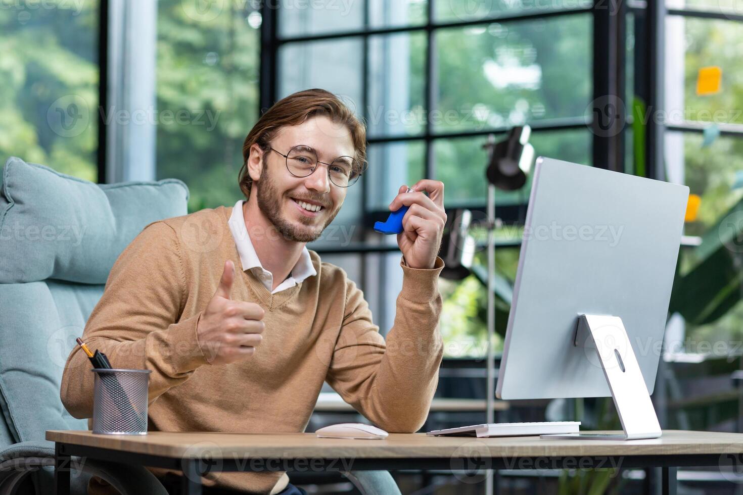 A young man poses for the camera, advertises a medicine for asthma and allergies. He sits in the office at a table with a computer, holds an inhaler in his hand, shows super with his hand, smiles. photo
