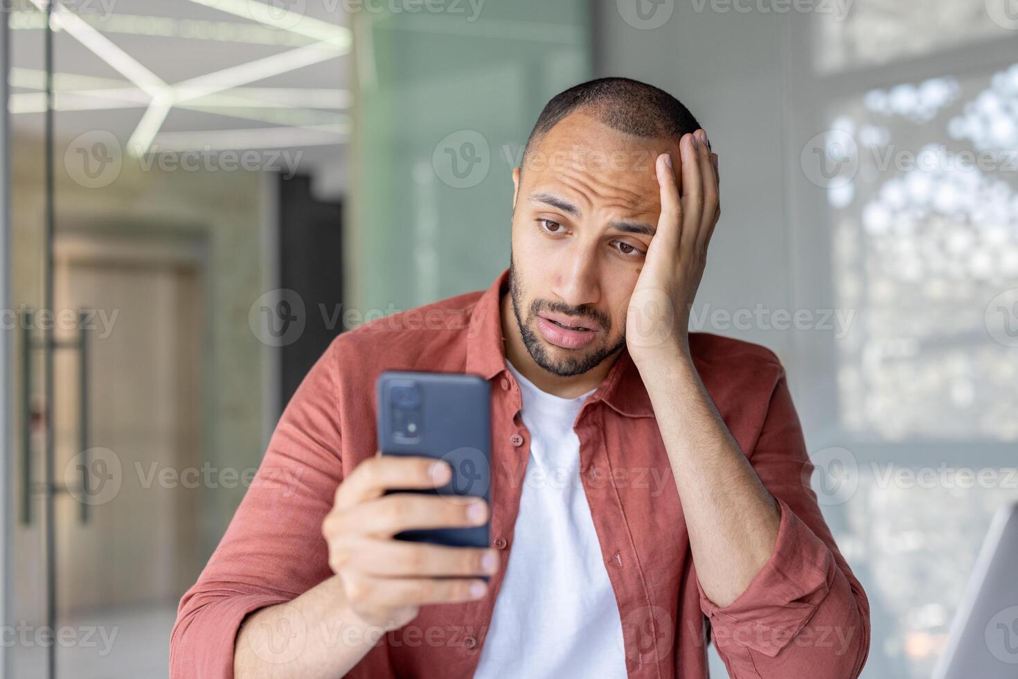 A man in an office looks at his phone with a worried expression, conveying anxiety, frustration, or critical thinking. The image portrays emotional stress connected to work or personal communication. photo