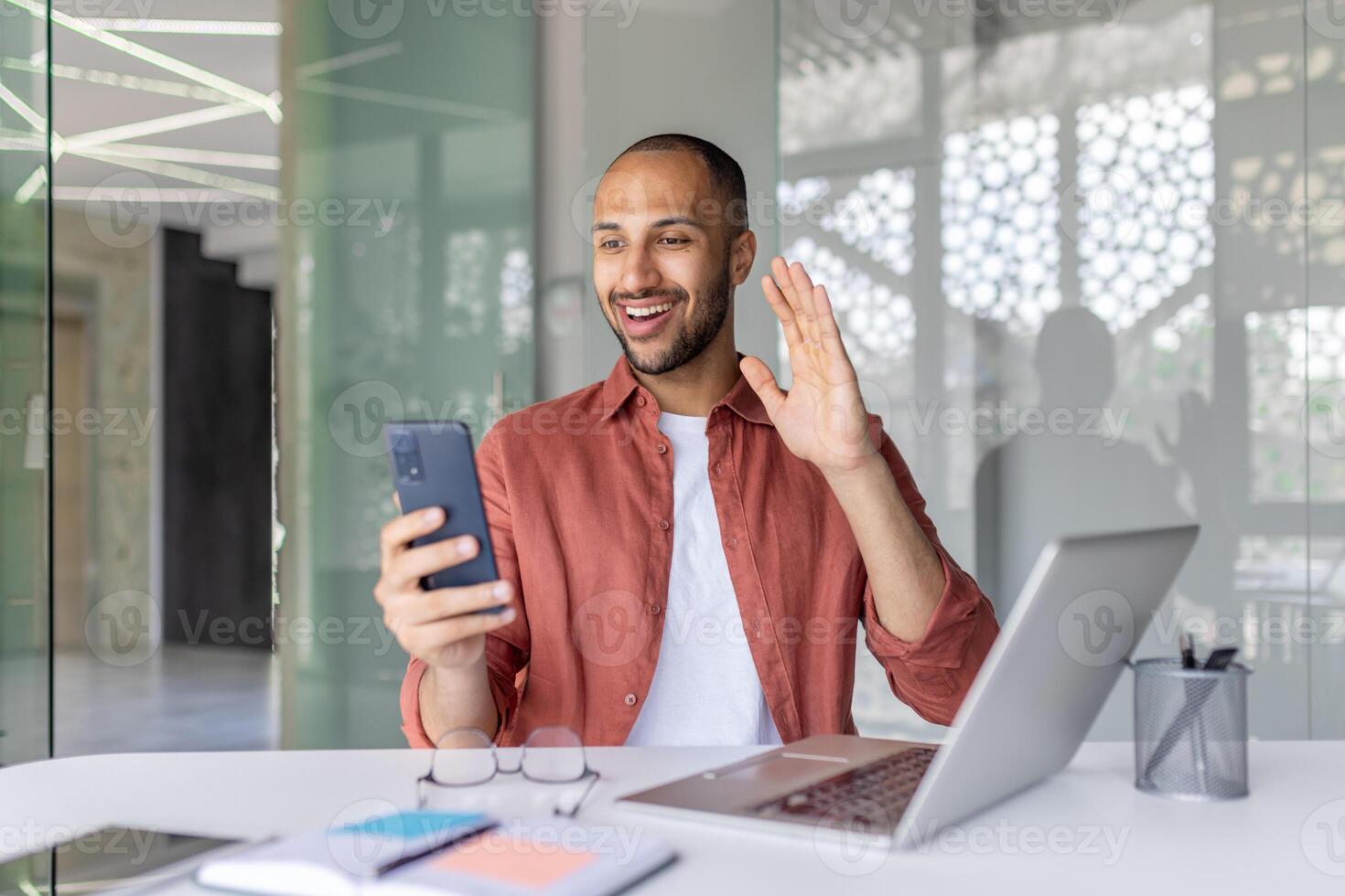 A cheerful businessman waves during a call in a contemporary office setting, using a smartphone and laptop. The casual environment highlights remote communication and connectivity. photo