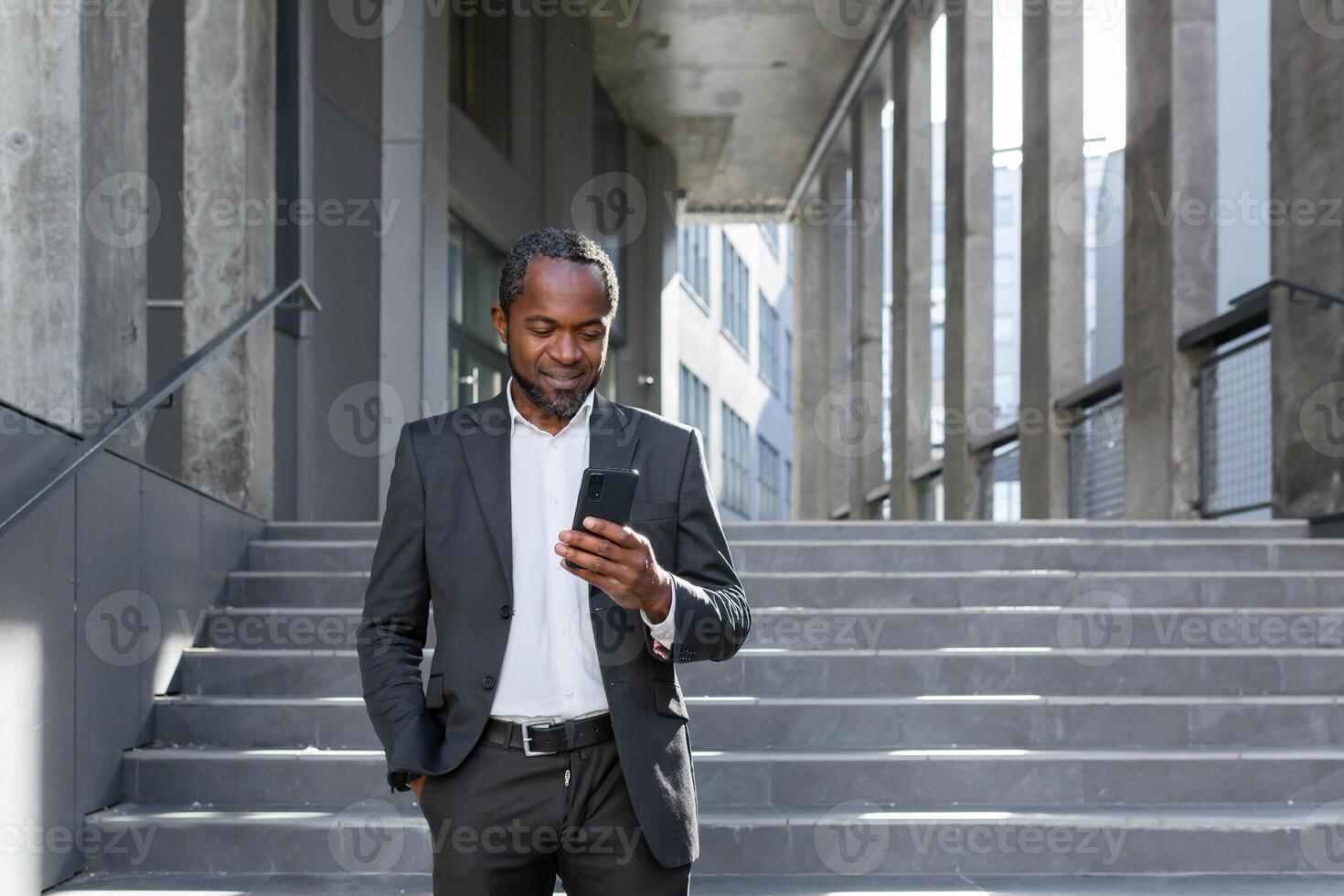 A successful mature African American man in a business suit is walking outside an office building, using a phone, typing a message and looking at the news online while smiling photo