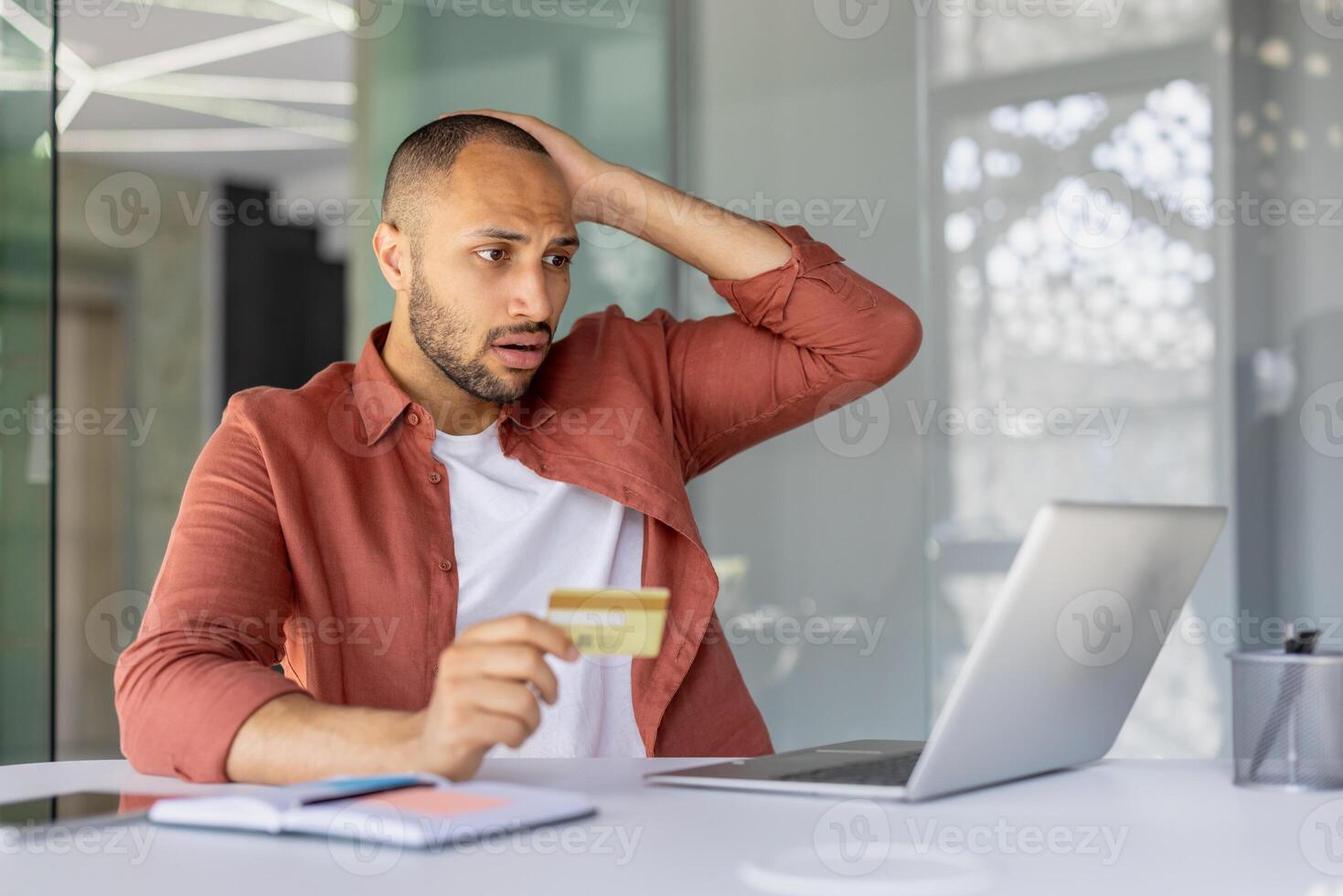 Man appears stressed holding credit card while checking laptop for payment issue or online banking problem in office. Concerned expression suggests financial trouble or online fraud worry. photo